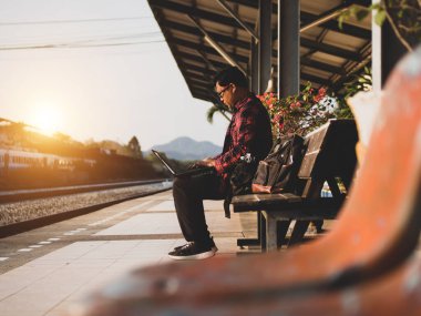 Asyalı adamın tren istasyonunda sosyal medyayı kullandığı yaz günü. Seyahat konsepti. Hipster adam telefon ve laptop kullanıyor sırt çantasıyla oturuyor ve trenin seyahat etmesini bekliyor..