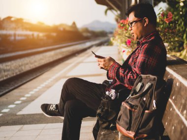 Asyalı adamın tren istasyonunda sosyal medyayı kullandığı yaz günü. Seyahat konsepti. Hipster adam telefon ve laptop kullanıyor sırt çantasıyla oturuyor ve trenin seyahat etmesini bekliyor..