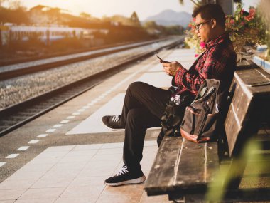 Asyalı adamın tren istasyonunda sosyal medyayı kullandığı yaz günü. Seyahat konsepti. Hipster adam telefon ve laptop kullanıyor sırt çantasıyla oturuyor ve trenin seyahat etmesini bekliyor..