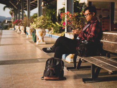 Asyalı adamın tren istasyonunda sosyal medyayı kullandığı yaz günü. Seyahat konsepti. Hipster adam telefon ve laptop kullanıyor sırt çantasıyla oturuyor ve trenin seyahat etmesini bekliyor..