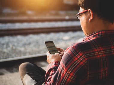Asyalı adamın tren istasyonunda sosyal medyayı kullandığı yaz günü. Seyahat konsepti. Hipster adam telefon ve laptop kullanıyor sırt çantasıyla oturuyor ve trenin seyahat etmesini bekliyor..