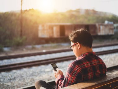 Asyalı adamın tren istasyonunda sosyal medyayı kullandığı yaz günü. Seyahat konsepti. Hipster adam telefon ve laptop kullanıyor sırt çantasıyla oturuyor ve trenin seyahat etmesini bekliyor..