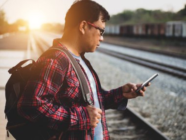 Asyalı adamın tren istasyonunda sosyal medyayı kullandığı yaz günü. Seyahat konsepti. Hipster adam telefon ve laptop kullanıyor sırt çantasıyla oturuyor ve trenin seyahat etmesini bekliyor..