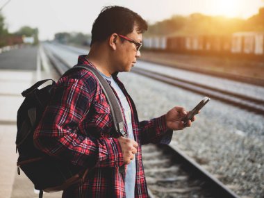 Asyalı adamın tren istasyonunda sosyal medyayı kullandığı yaz günü. Seyahat konsepti. Hipster adam telefon ve laptop kullanıyor sırt çantasıyla oturuyor ve trenin seyahat etmesini bekliyor..
