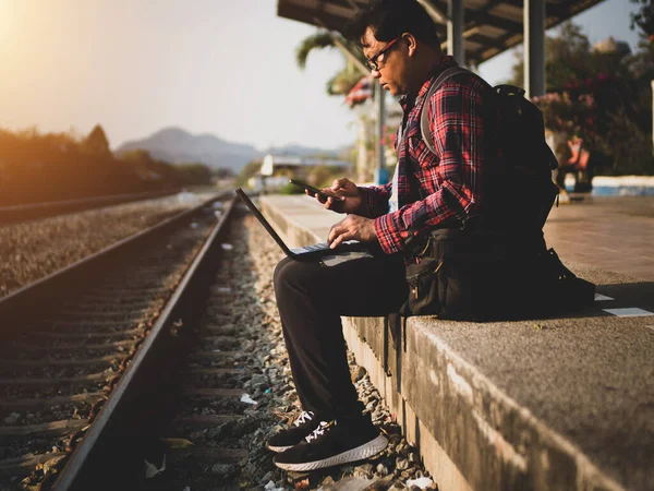 Asyalı adamın tren istasyonunda sosyal medyayı kullandığı yaz günü. Seyahat konsepti. Hipster adam telefon ve laptop kullanıyor sırt çantasıyla oturuyor ve trenin seyahat etmesini bekliyor..