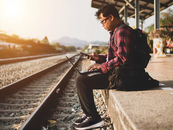 Asyalı adamın tren istasyonunda sosyal medyayı kullandığı yaz günü. Seyahat konsepti. Hipster adam telefon ve laptop kullanıyor sırt çantasıyla oturuyor ve trenin seyahat etmesini bekliyor..