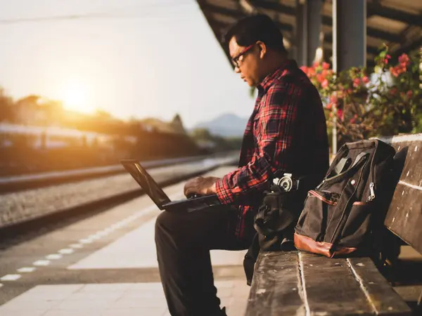 Asyalı adamın tren istasyonunda sosyal medyayı kullandığı yaz günü. Seyahat konsepti. Hipster adam telefon ve laptop kullanıyor sırt çantasıyla oturuyor ve trenin seyahat etmesini bekliyor..