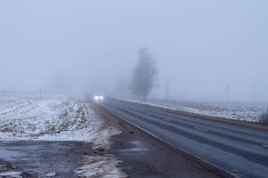 Işıklar açıkken araba sonbahar sabahının erken saatlerinde siste gider.