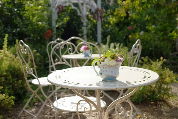 A white coffee tables with chairs stands in the garden among green bushes. Decoration from flowers in vase. Beautifully vintage decorated garden for a banquet.