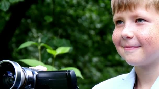 Niño con cámara de vídeo filma película sobre la naturaleza del fondo verde del parque . — Vídeos de Stock