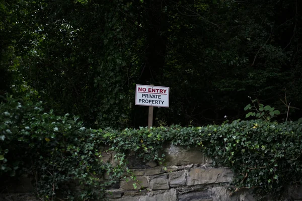 No Entry sign / Private Property red on white, attached to a wooden post behind an old stone wall covered with ivy.