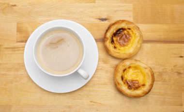 Kahve fincanı ve pastel de nata olarak bilinen Portekiz kremalı tartları. Tahta masanın üstünden aşağı doğru..