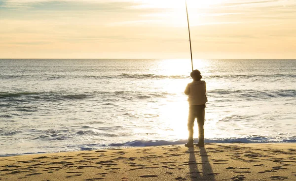 Elderly man fishing, in silhouette at sunset on beach