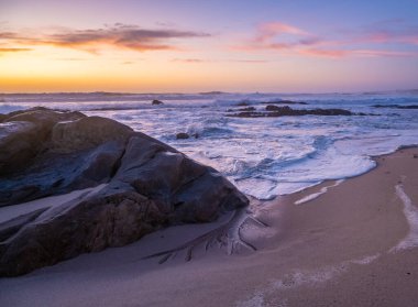 Alacakaranlıkta sahilin üzerinde geniş açılı çekim. Canlı turuncu, dalgalı dalgalar ve solunda büyük koyu bir kaya var..