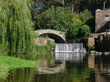 Beautiful summer scene on Este river in Portugal with ancient stone bridge, waterfall and overhanging willow tree. clipart