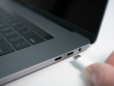 Elle beyaz bir USB-C / Tip C veya Thunderbolt 3 kablosunu gri bir dizüstü bilgisayar / dizüstü bilgisayara bağlamak.