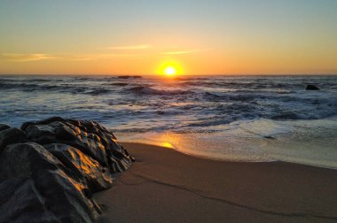 Gün batımında plajda alçak açılı çekim. Suyun üzerinde turuncu yansımalar ve solda ıslak kum büyüklüğünde kaya var.