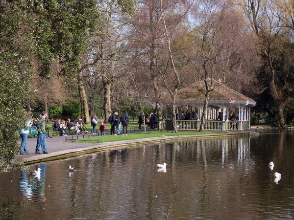 St. Stephen 's Green Park in Dublin, Ireland during autumn, looking over lake and bandstand as people walk by
