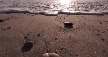 Yazın kumsalda kum ve çakıl taşlarının üzerinde dalgalar. Alçak açı yakın çekim, kamera yere yakın geri hareket ediyor..