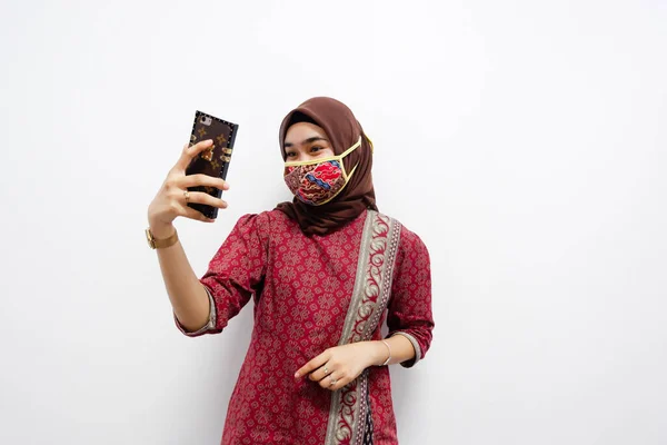 Young beautiful Asian woman that wears hijab and batik is doing some selfie pose on white background. And also she wears batik-pattern mask to protect her from covid-19
