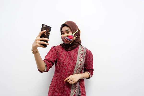 Başörtüsü takan genç ve güzel Asyalı kadın beyaz arka planda selfie pozu veriyor. Ve ayrıca onu covid-19 'dan korumak için batik desenli maske takıyor.