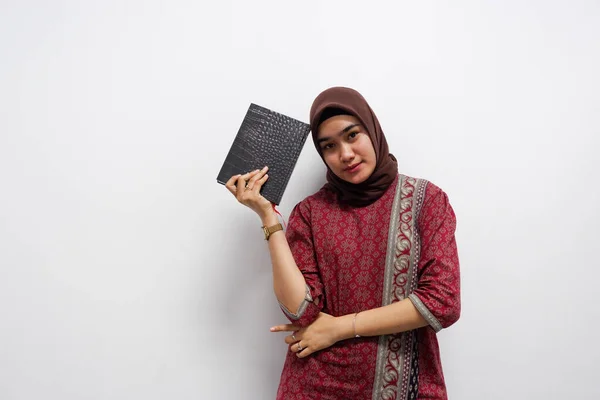 Genç Asyalı kadın bir kitap tutuyor ve bir şey düşünürken kameraya bakıyor. Genç Asyalı öğrenci batik üniforma giyer ve beyaz arka plana tesettür takar..