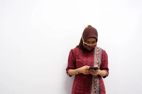 Genç Asyalı kadın telefonuna bakıyor. Beyaz arkaplana batik desenli maske takmak.