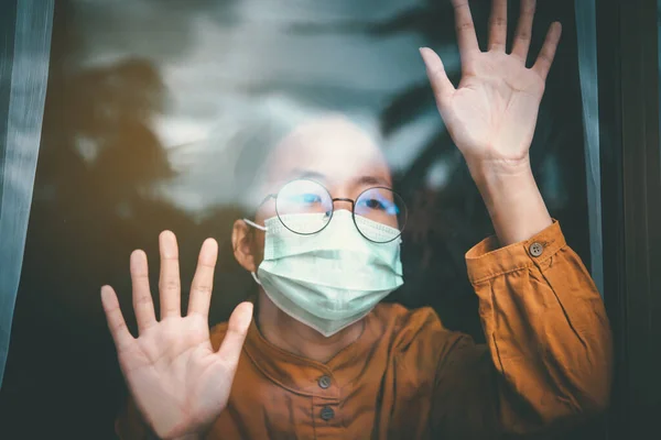 Asian girl wearing glasses, wearing a mask, looking out the window, is bored of having to detain and treat the illness at home alone. Concept home quarantine, prevention COVID-19