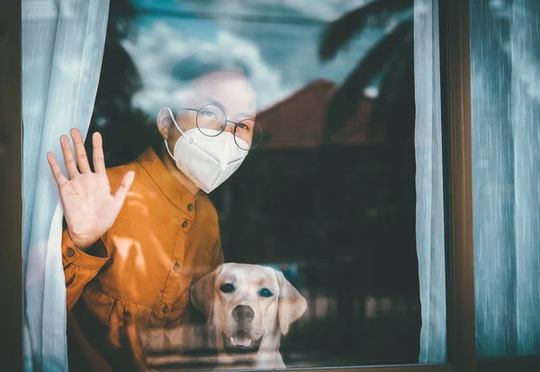 Asian girl wearing glasses, wearing a mask, with Labrador dog looking out the window, is bored of having to detain and treat the illness at home alone. Concept home quarantine, prevention COVID-19