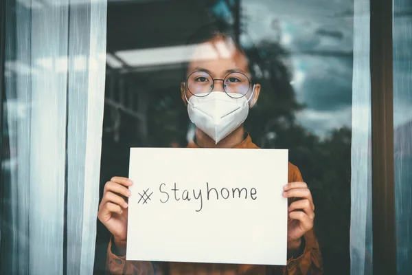 Asian girl wearing glasses, wearing a mask, holding paper labeled #stayhome, is bored of having to detain and treat the illness at home alone. Concept home quarantine, prevention COVID-19