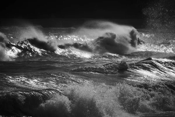 A stormy sea with gale force winds and the high tide creates huge waves which crash on the rocky beach. The setting sun glints across the water and the spray creates a mist across the water.