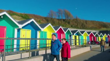 Barry, Glamorgan / Galler Vadisi - 20 Ocak 2020: Güneşli bir kış öğleden sonrasında, sıcak paltolu insanlar Güney Galler 'deki Barry Adası' ndaki parlak renkli sahil kulübelerinin yanında geziniyorlar.. 