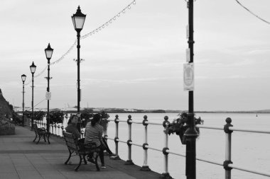 Güneş battı ve insanlar Penarth gezinti güvertesi ve iskele boyunca akşam yürüyüşünün tadını çıkarıyor. Viktorya dönemi pavyonu bu sahil kasabasında çok iyi bilinen bir simgedir ve insanlar tahta kaldırım boyunca yürümeyi severler.