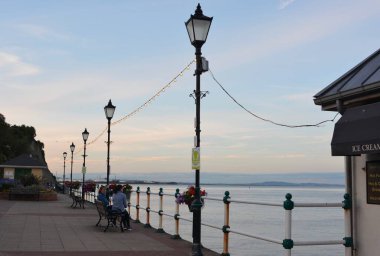 Güneş battı ve insanlar Penarth gezinti güvertesi ve iskele boyunca akşam yürüyüşünün tadını çıkarıyor. Viktorya dönemi pavyonu bu sahil kasabasında çok iyi bilinen bir simgedir ve insanlar tahta kaldırım boyunca yürümeyi severler.