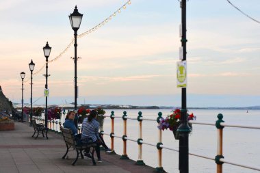 Güneş battı ve insanlar Penarth gezinti güvertesi ve iskele boyunca akşam yürüyüşünün tadını çıkarıyor. Viktorya dönemi pavyonu bu sahil kasabasında çok iyi bilinen bir simgedir ve insanlar tahta kaldırım boyunca yürümeyi severler.
