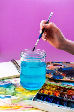 Suluboya resim, mavi suyla konserve, fırça ve boya. yazıtlar, yaratıcılık konsepti ve eflatun arkaplan üzerindeki sanat için