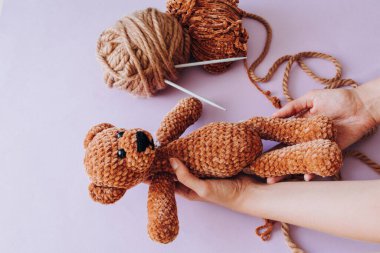 Kahverengi örgü ipliği. Örgü iğneleri ipliğe sıkıştı. Eflatun bir arka planda dişi ellerinde örülmüş kahverengi ayı. Hobi ve doldurulmuş oyuncak konsepti. Metin ve ekleme için yer. 