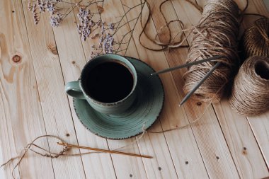 Örgü örmek ve örgü örmek için iplik. Örgü iğneleri ipliğe saplandı, örgü kancası örmeye başladı. Yanındaki çay tabağının üzerinde kahve ile birlikte yeşil bir fincan. Ahşap bir arka planda. Hobi konsepti.