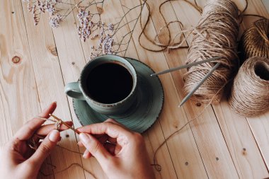 Örgü örmek ve örgü örmek için iplik. İpliğe saplanmış örgü iğneleri. Kadın elleri örgü örmeye başladı. Yanındaki çay tabağının üzerinde kahve ile birlikte yeşil bir fincan. Ahşap bir arka planda. Hobi ve eğlence konsepti.