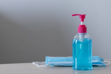 Mavi alkol jeli şişesi maskeyle birlikte sağ tarafa yerleştiriliyor. Masada ve beyaz duvarda Alkol El anti-virüs ve anti-virüs Covid-19 'u yıkıyor.