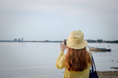 Kadın arkada duruyor ve el kamerasıyla deniz sahilindeki günbatımını çekiyor. Balıkçı teknelerinin açık havada hayatta kaldığı sahil kıyısı boyunca..