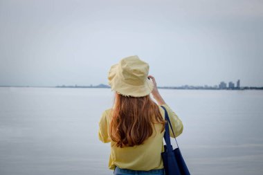 Kadın arkada duruyor ve el kamerasıyla deniz sahilindeki günbatımını çekiyor. Balıkçı teknelerinin açık havada hayatta kaldığı sahil kıyısı boyunca..