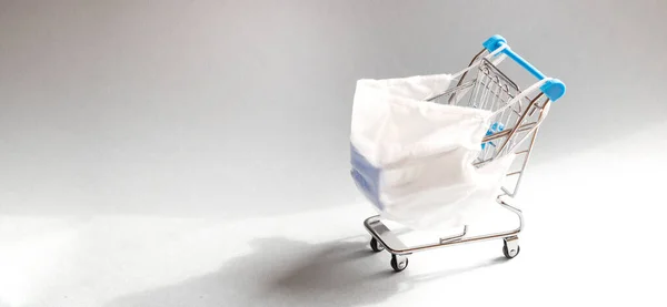 Shopping cart with protective medical mask against coronavirus. Safe and online shopping on quarantine concept. Light blue background, copy space.