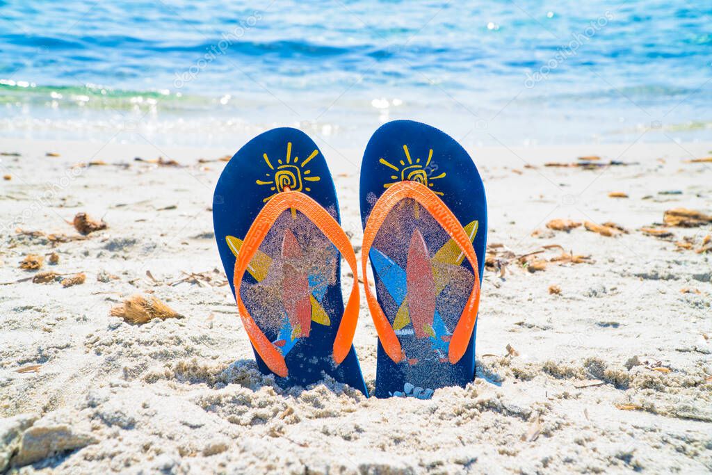 Blue and orange flip flops on the shore of a blue and crystalline sea during the summer holidays. Ideal for advertising travel, holidays and seaside resorts