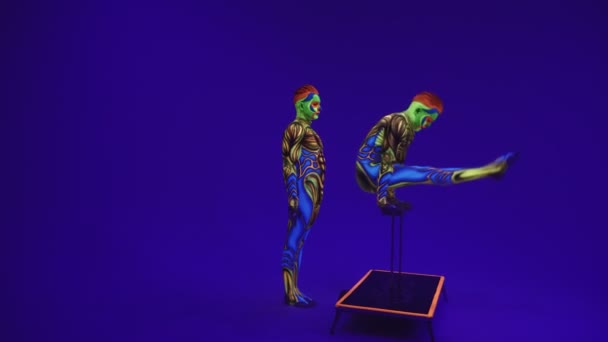 ネオンコスチュメストのアクロバティックな双子は 手と頭の上でスタンドを実行します エイリアンのスタイルで紫外線メイクで青いサイクロラマに — ストック動画