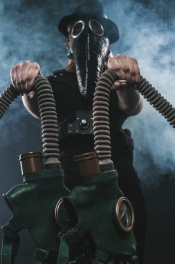 Veba maskeli ve siyah şapkalı bir kızın elinde gaz maskeleri var. dumanın arka planına karşı