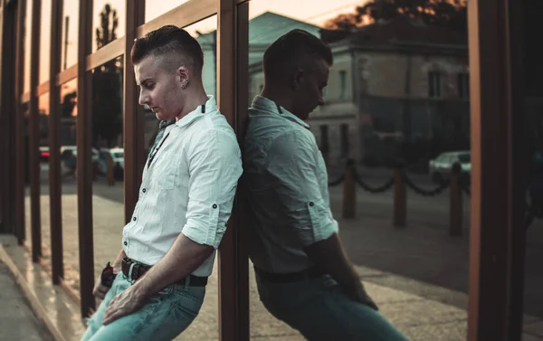 a guy in a white shirt and blue jeans is leaning against a glass wall. reflection