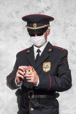 Üniformalı, polis rozetli, siyah kılıflı, beyaz tıbbi maskeli ve siyah gözlüklü bir Rus polis teğmeni Rubik Küpü 'nü bir araya getiriyor.