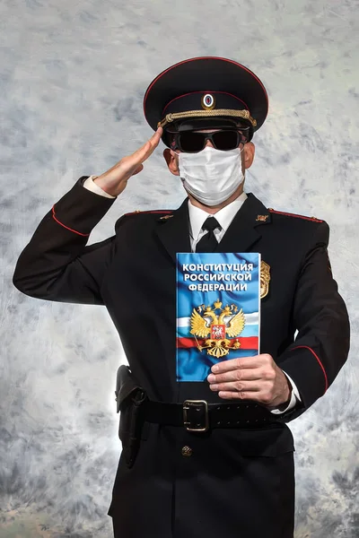 Russian police officer lieutenant in a white medical mask and black glasses holds the Constitution of the Russian Federation on which is written \
