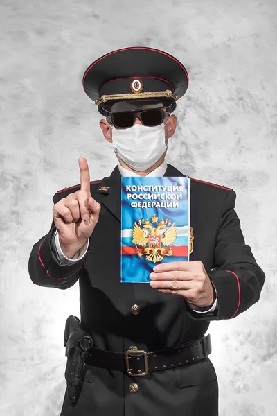Russian police officer in uniform in a white medical mask and black glasses holds the index finger up and holds the Constitution on which is written in russian \
